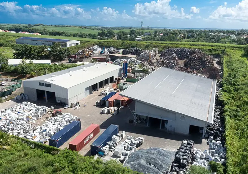 Vue aérienne d'un site de recyclage avec deux hangars à toit gris, conteneurs bleus et rouges, stocks de matériaux et ferraille, entouré de végétation sous un ciel bleu.