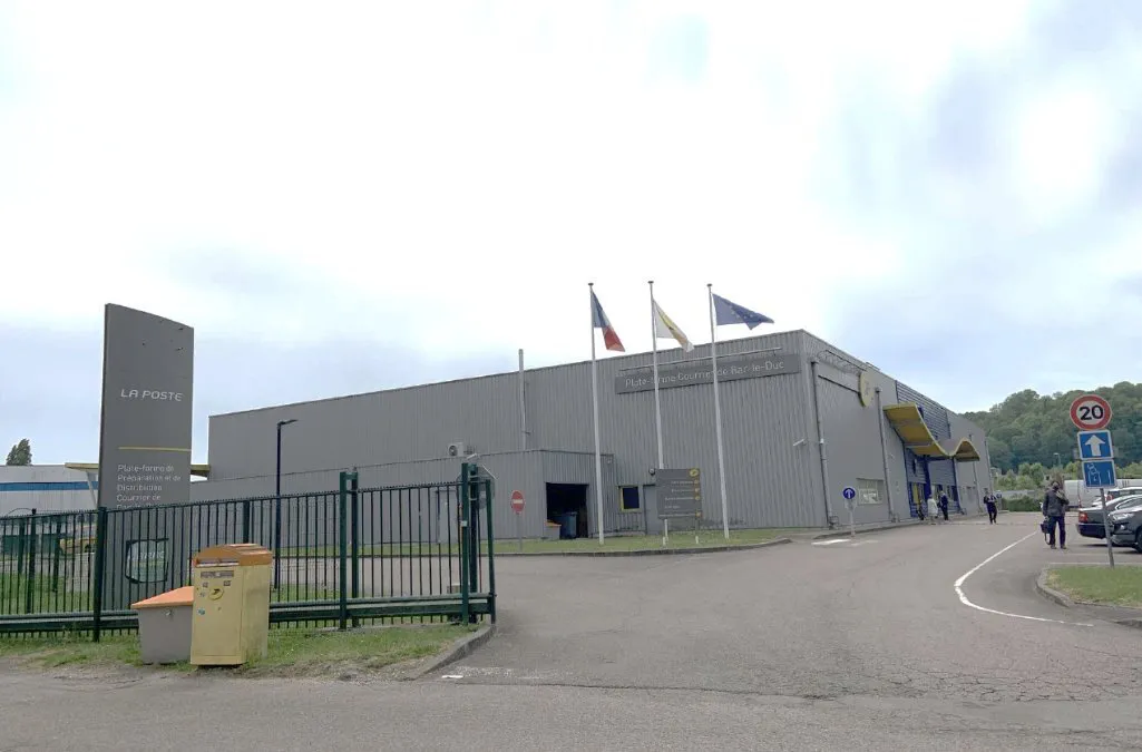 Bâtiment d'activités en bardage gris à Bar-le-Duc, avec trois mâts portant des drapeaux, un totem d'entrée, une clôture verte et une voie d'accès goudronnée sous un ciel nuageux.